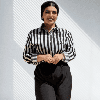 Woman wearing a black and white striped shirt and black pants, smiling while standing in a well-lit area.