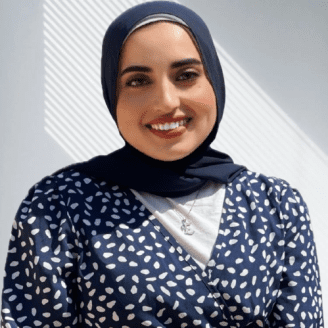Woman wearing a navy blue hijab and a patterned blouse, smiling at the camera with soft lighting.