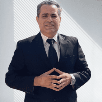 Man in a black suit and tie standing with hands clasped, wearing a watch, against a light background.