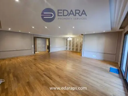 Spacious empty room with wooden flooring and neutral-colored walls, featuring recessed lighting and a chandelier.