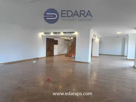 Spacious interior of an empty room with wooden flooring and a staircase in the background.