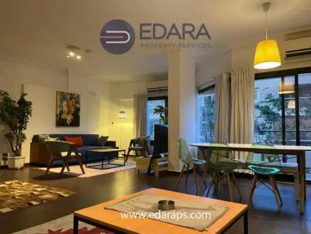 Modern living room with a blue sofa, wooden dining table, and decorative plants in a well-lit space.