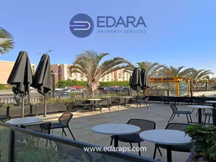 Outdoor seating area with tables and chairs under umbrellas, featuring palm trees in the background.