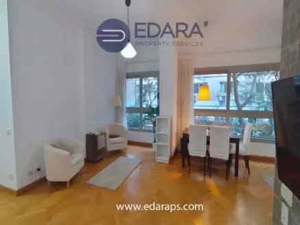 Modern living room with a dining area, featuring two armchairs, a dining table, and large windows.