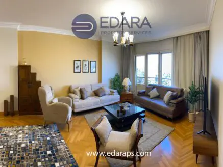 Modern living room with beige sofas, a coffee table, and a patterned rug, featuring large windows and decorative elements.