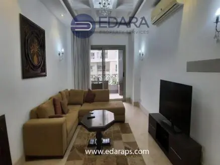 Living room with a beige sectional sofa, coffee table, and a television against a light-colored wall.