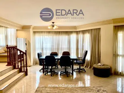 Spacious living area with a large table and chairs, featuring a staircase and curtains in a well-lit room.