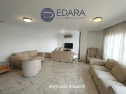 Living room featuring beige sofas arranged around a marble floor with natural light from large windows.
