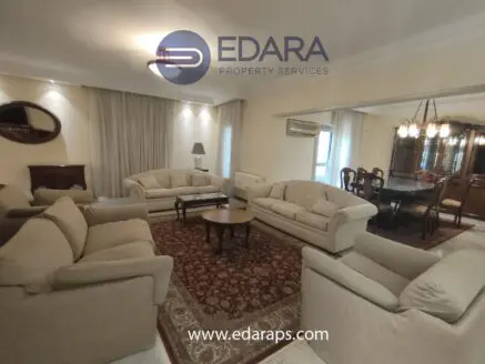 Spacious living room featuring beige sofas arranged around a coffee table with a dining area in the background.