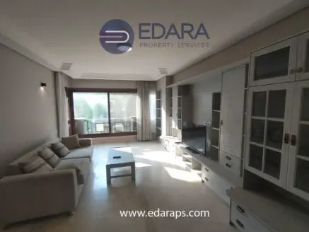 Spacious modern living room with a sofa, TV unit, and large windows allowing natural light.