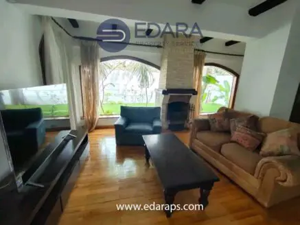 Living room featuring a sofa, armchair, coffee table, and television against a backdrop of large windows.