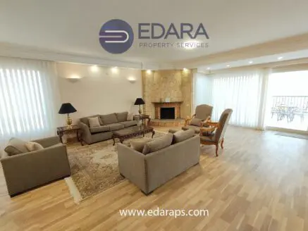 Living room featuring beige sofas, wooden furniture, and a stone fireplace with large windows for natural light.