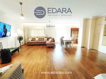 Bright living room featuring a large sofa, wooden flooring, and a television mounted on the wall.