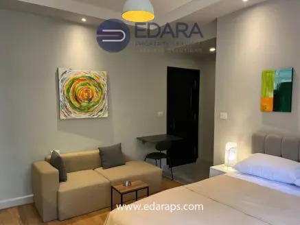 Contemporary living room featuring a beige sofa, abstract artwork, and a bed in a neutral color scheme.