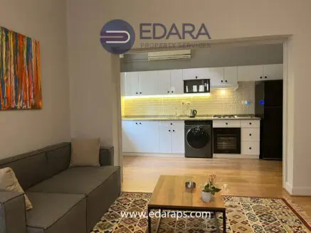 Contemporary living room featuring a gray sofa and a view of a kitchen with white cabinets and appliances.