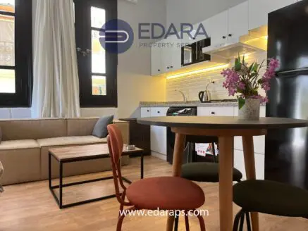 Interior view of a modern apartment featuring a kitchen, dining table, and seating area with a sofa and decorative flowers.