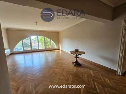 Spacious empty room featuring large arched windows and wooden flooring, with a small table in the center.