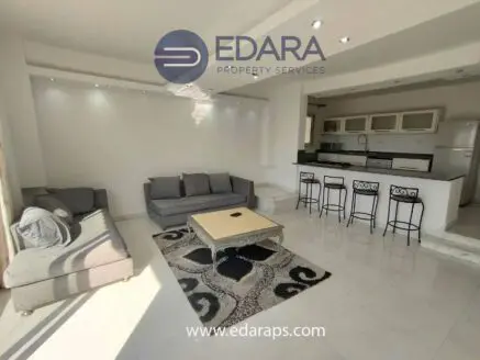Contemporary living room with a gray sofa and a kitchen area featuring bar stools and a dining table.