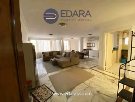 Bright living room featuring a large sofa set and an open kitchen area with marble flooring.
