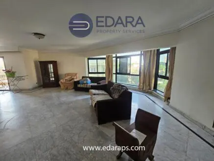 Spacious living room featuring large windows, a sofa, and a wooden cabinet in a neutral color scheme.