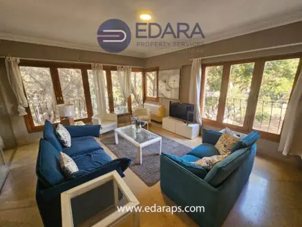 Spacious living room featuring a blue sofa set, coffee table, and large windows allowing natural light.