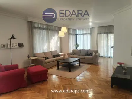 Spacious living room featuring light-colored sofas, a coffee table, and large windows with sheer curtains.