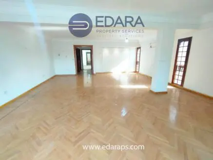Spacious empty room with wooden herringbone flooring and large windows allowing natural light.