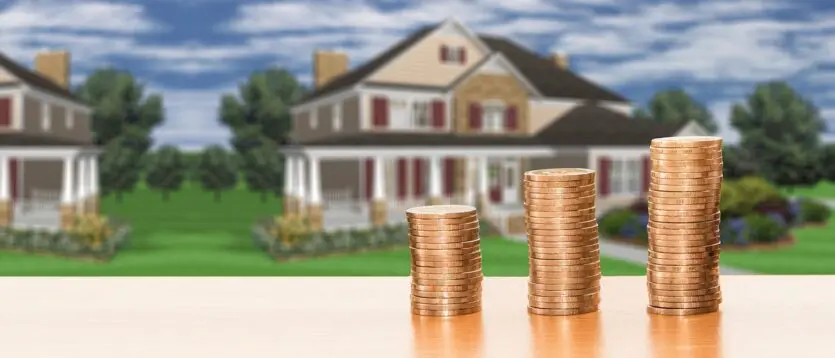 Four stacks of coins of varying heights positioned on a table with a house in the background.
