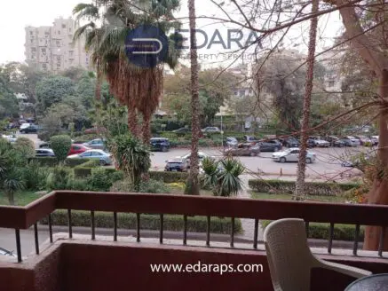 Balcony view showing a garden area with palm trees and parked cars in the background.