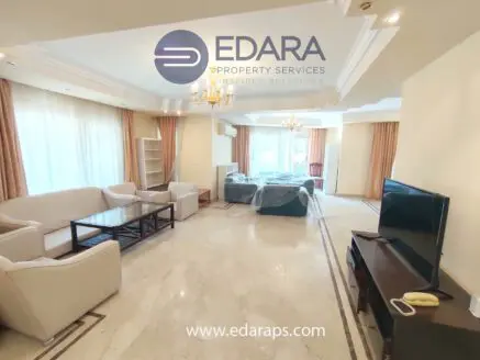 Spacious living room featuring a white sofa, coffee table, and television in a well-lit environment.