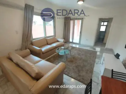 Living room featuring two beige sofas, a glass coffee table, and large windows allowing natural light.