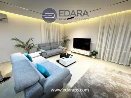 Modern living room featuring gray sofas, a white TV unit, and indoor plants in a well-lit space.