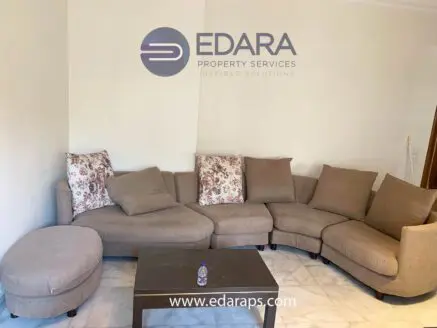 L-shaped beige sofa with floral decorative cushions and a small round ottoman in a neutral setting.