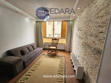 Living room featuring a gray sofa, a rocking chair, and a stone accent wall with natural lighting.