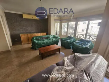 Living room featuring furniture covered in plastic, including a sofa and coffee table, with a light-colored wall and flooring.