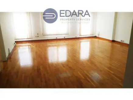 Spacious empty room with polished wooden flooring and large windows allowing natural light.