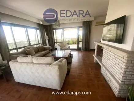Spacious living room featuring a light-colored sofa set and a wooden coffee table with large windows allowing natural light.