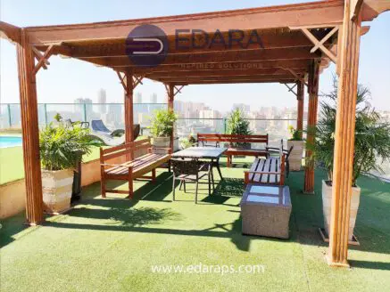 Rooftop seating area featuring a wooden pergola, tables, and benches surrounded by greenery.