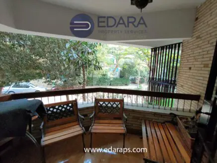 Wooden benches and chairs arranged on a balcony with a view of greenery and a street.