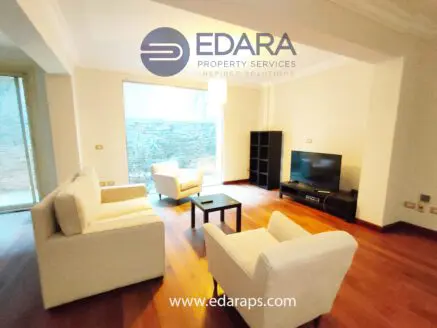 Bright living room featuring white sofas, a television, and wooden flooring.