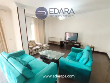 Living room featuring teal sofas, a glass coffee table, and a television set against a neutral wall.