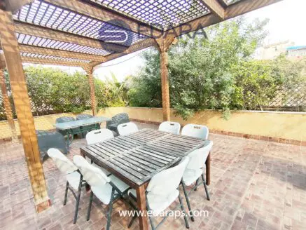 Wooden dining table surrounded by white chairs under a wooden pergola in an outdoor setting.