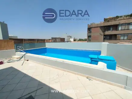 Rooftop swimming pool with a blue surface and tiled deck, featuring a ladder and seating area.