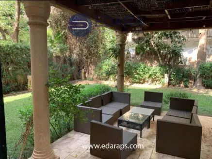 Outdoor seating area featuring rattan furniture arranged around a glass coffee table in a garden setting.