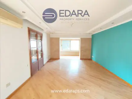 Interior view of a room featuring a blue accent wall and wooden flooring, with large windows allowing natural light.