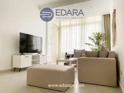 A modern living room featuring a beige sofa, television, and decorative plant in a bright space.