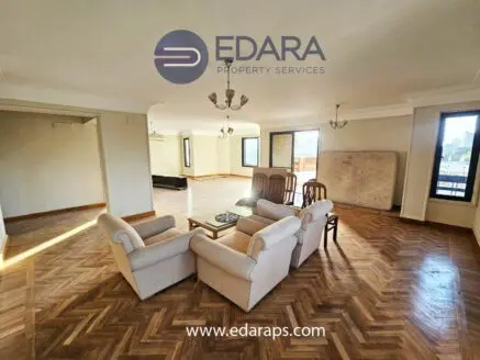 Spacious living room featuring light-colored armchairs and wooden herringbone flooring with large windows.