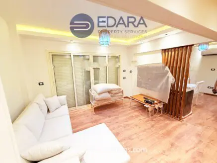 Contemporary living room with a light-colored sofa and wooden flooring, featuring a dining area and large windows.