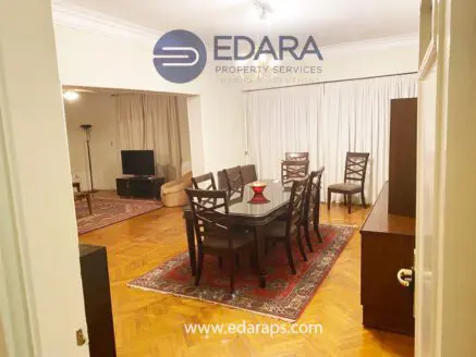 Interior view of a living room featuring a dining table and chairs, with a sofa and television in the background.