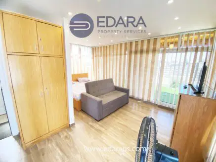 Furnished apartment interior with a sofa bed, wooden cabinets, and large windows with blinds.